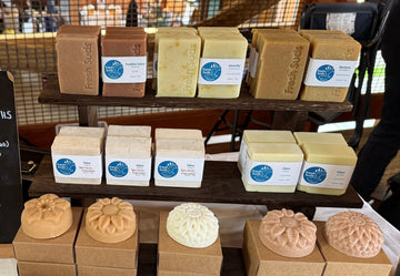 Fresh Suds handmade soap bars on display at a market — rectangular olive, calendula, dandelion, and rosehip bars alongside floral-molded seasonal bars, each with their scent and skin-type label.